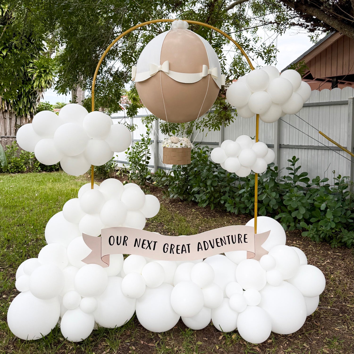Gender Reveal Hot Air Balloon display featuring a beige and white jumbo balloon with paper bows, a basket filled with flowers, and a banner reading “Our Next Great Adventure,” surrounded by white balloon clouds, perfect for baby showers, gender reveal parties, and adventure-themed celebrations.