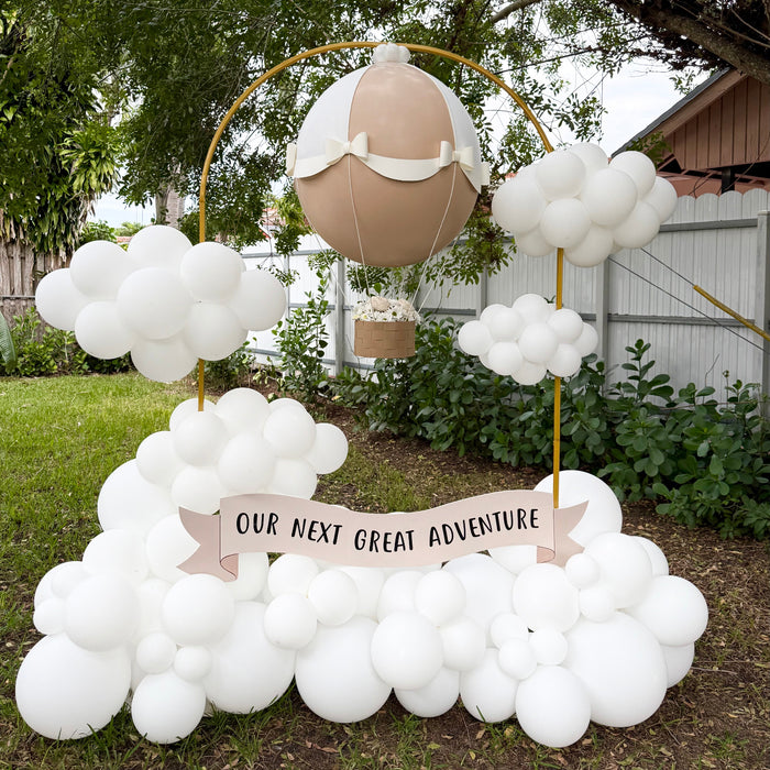 Gender Reveal Hot Air Balloon display featuring a beige and white jumbo balloon with paper bows, a basket filled with flowers, and a banner reading “Our Next Great Adventure,” surrounded by white balloon clouds, perfect for baby showers, gender reveal parties, and adventure-themed celebrations.