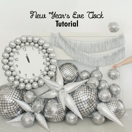 Glamorous New Year’s Eve balloon clock display featuring a large silver balloon clock with bold black hands pointing to midnight, surrounded by reflective disco ball balloons, metallic balloon stars, and silver fringe. Perfect for party decor or photo booth backdrops. Inspired by Lilly Jimenez’s tutorial for festive balloon installations and NYE celebration ideas.