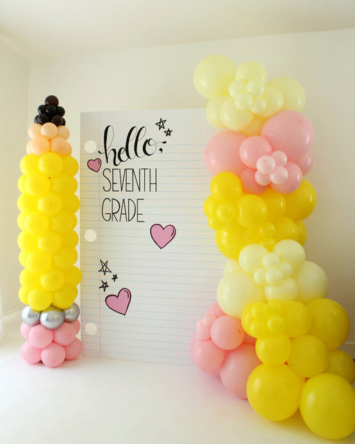 Large 48"x72" notebook paper backdrop reading “Hello, Seventh Grade” with pink heart and star sketches, flanked by colorful balloon columns shaped like a pencil and flowers. Perfect for first day of school photo ops, classroom decorations, and teacher events. Includes customizable grades from Pre-K to Eighth Grade and matching SVG files.