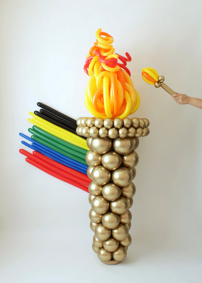 Large Olympic-themed balloon torch made with gold balloons and topped with flame-like balloons in red, orange, and yellow. Colorful balloon “rings” representing Olympic colors extend from the back. Ideal for Olympic party decorations, sports events, and school spirit displays.