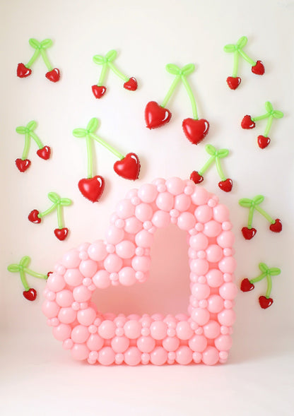 Pink open heart balloon sculpture surrounded by heart-shaped balloon cherries with green stems, creating a whimsical Valentine's display.