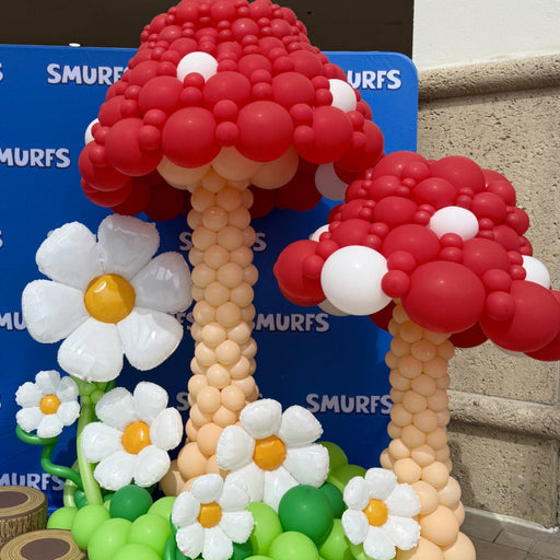 Oversized red balloon mushrooms with white polka dots, balloon daisies, and green balloon grass in front of a blue themed backdrop, perfect for whimsical party decor, themed events, photo backdrops, and playful balloon installation inspiration.