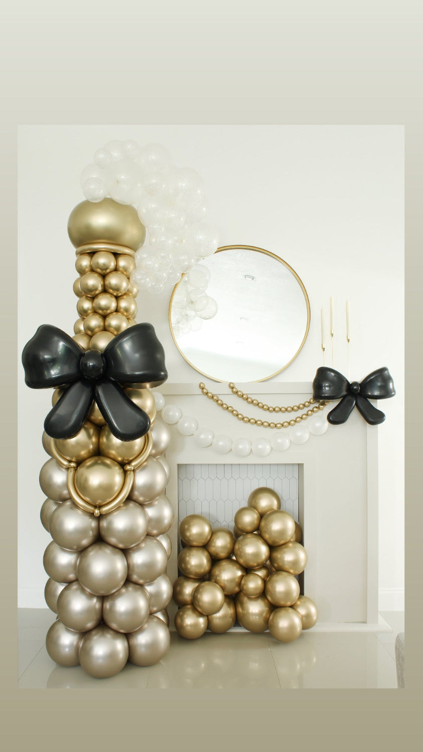 Oversized champagne bottle made entirely from metallic gold and pearl balloons, featuring a large black balloon bow. Styled beside a white fireplace with matching balloon decor, perfect for weddings, New Year's Eve, and cocktail hour photo ops. Designed by Lilly Jimenez.
