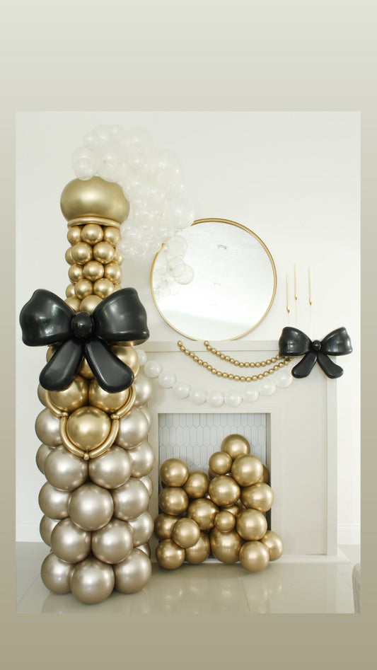 Oversized champagne bottle made entirely from metallic gold and pearl balloons, featuring a large black balloon bow. Styled beside a white fireplace with matching balloon decor, perfect for weddings, New Year's Eve, and cocktail hour photo ops. Designed by Lilly Jimenez.