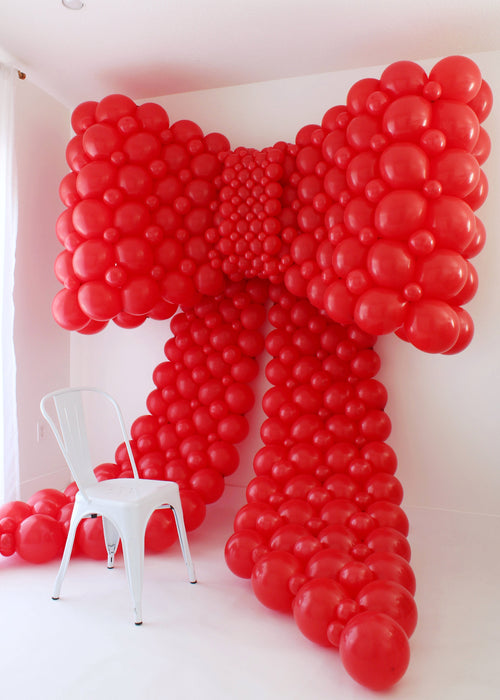 Giant red linking balloon bow made with rounded bubble-style clusters, displayed in a white studio with a white metal chair for scale. The bow features large loops and long tails, showing the dramatic size students can create in the Linking Balloon Bows Class.