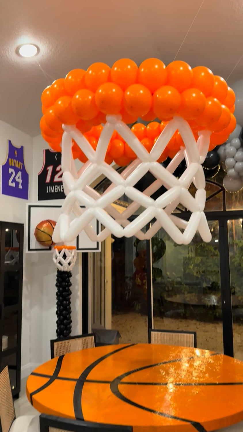 Basketball-themed party setup featuring a giant balloon basketball hoop suspended above a round table decorated like a basketball court. Includes balloon jersey wall decor, backboard props, and coordinating black, gray, and orange balloon elements. Perfect for sports-themed birthdays and DIY party decorators using balloon sculpture techniques.