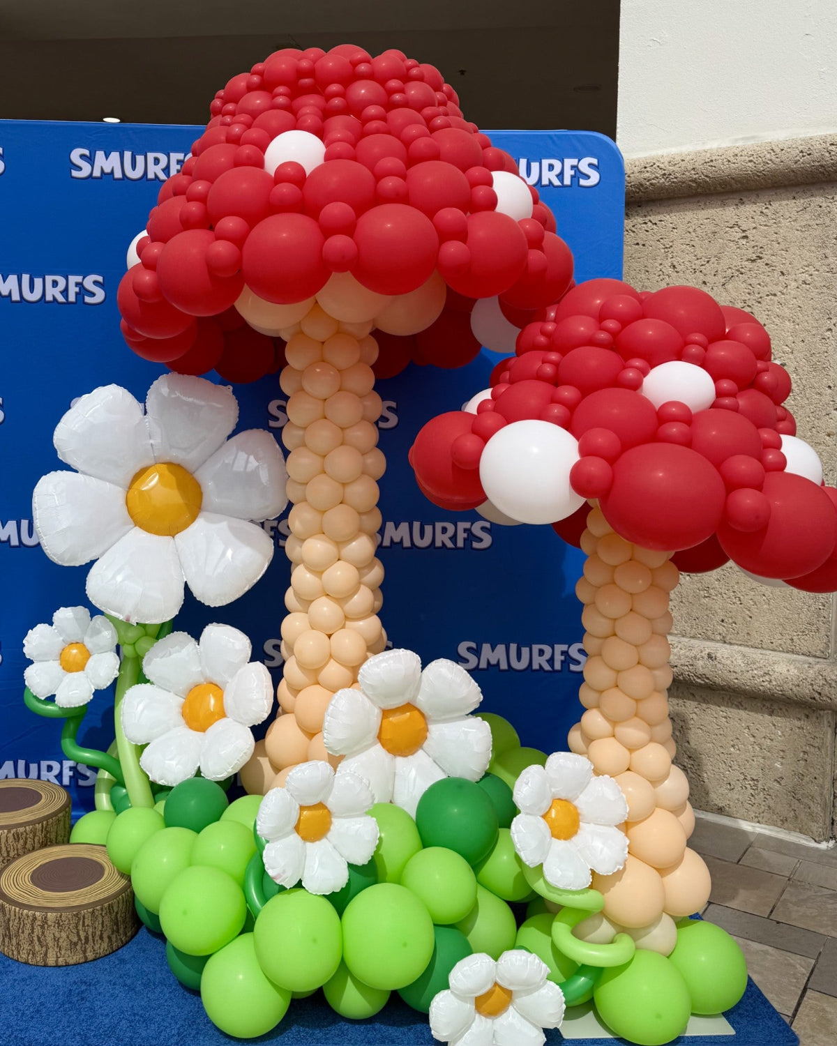 Whimsical balloon display featuring two oversized red mushroom sculptures with blush stalks, surrounded by white balloon daisies with yellow centers and bright green balloon grass. Set against a Smurfs-branded backdrop, this playful scene was created as part of The Creative Heart Studio’s Mushroom Balloon Class & Recipes—perfect for themed parties, brand activations, and immersive balloon decor.