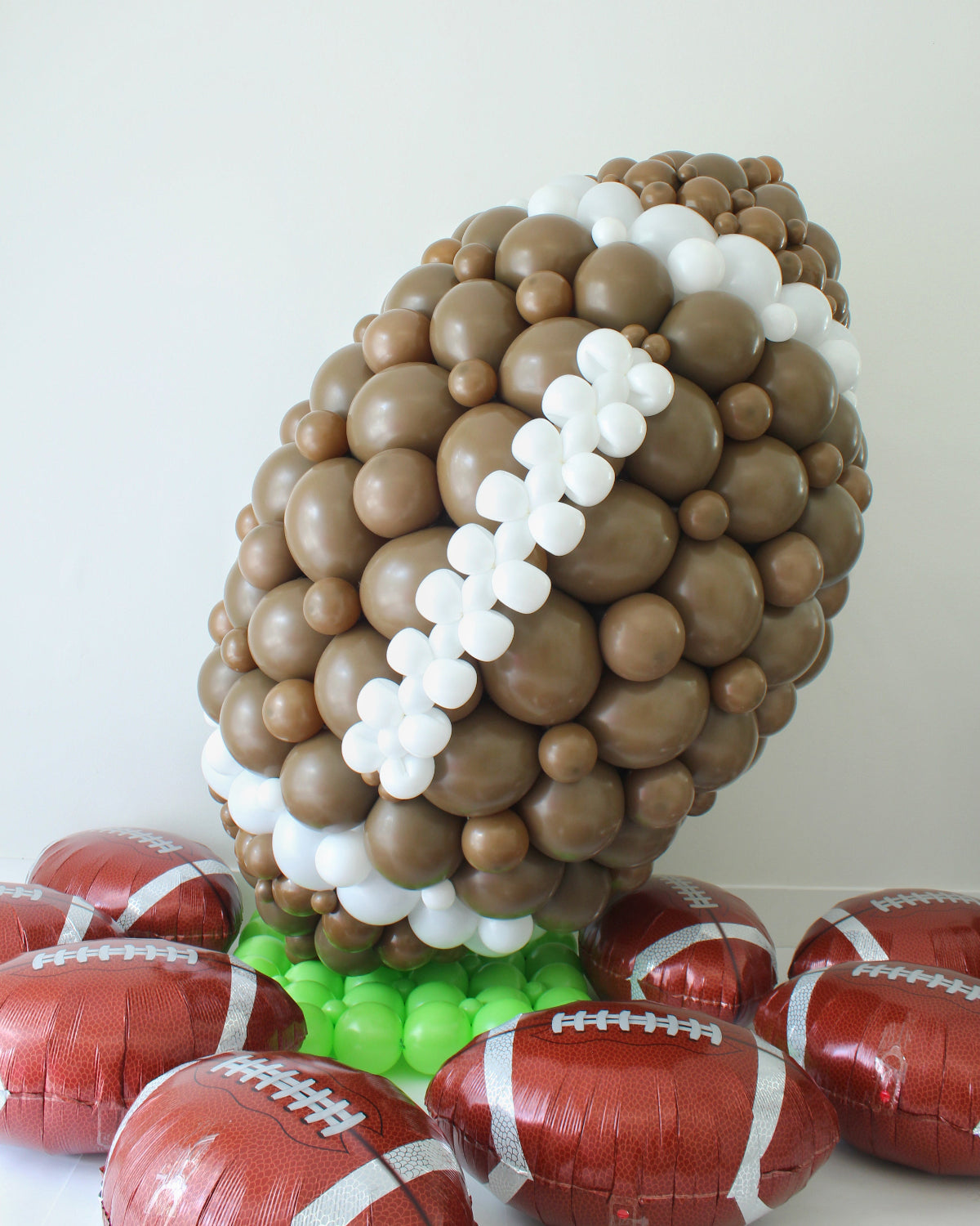 Large football balloon sculpture created with brown, white, and green balloons, displayed on a base of foil football balloons. Perfect for Super Bowl parties, tailgate events, sports banquets, and school spirit celebrations, this creative balloon design makes a bold and playful statement for any football-themed event.