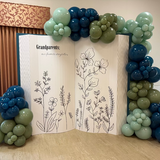Open Book Backdrop decorated with clusters of green and blue balloons, perfect for storybook-themed events, photo booths, graduations, and weddings. The oversized foamboard book features illustrated floral designs and customizable inside pages, making it a versatile digital download template for event decorators and balloon stylists.