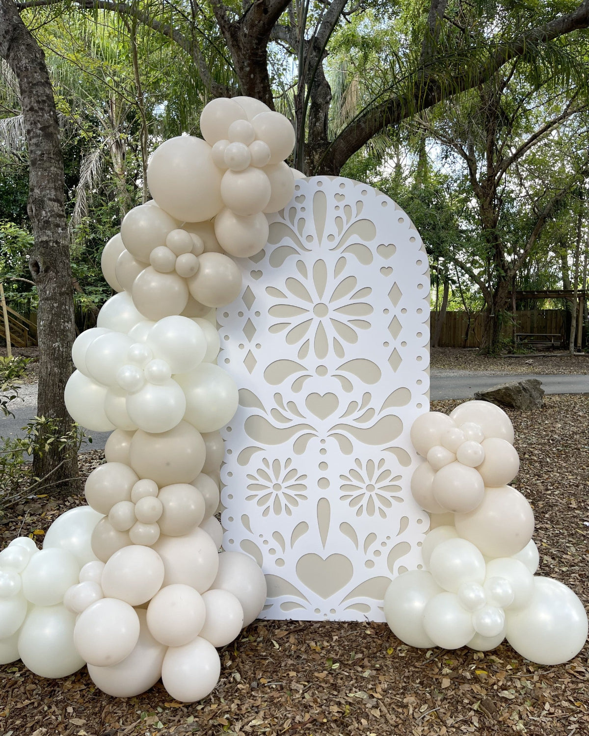 Large white papel picado-style foam board backdrop decorated with cream and ivory balloon clusters. Displayed outdoors among trees, this elegant party setup is ideal for weddings, baby showers, birthdays, or Día de Muertos events. Design made using a laser cut or CNC machine with files available in SVG, DXF, and printable formats.