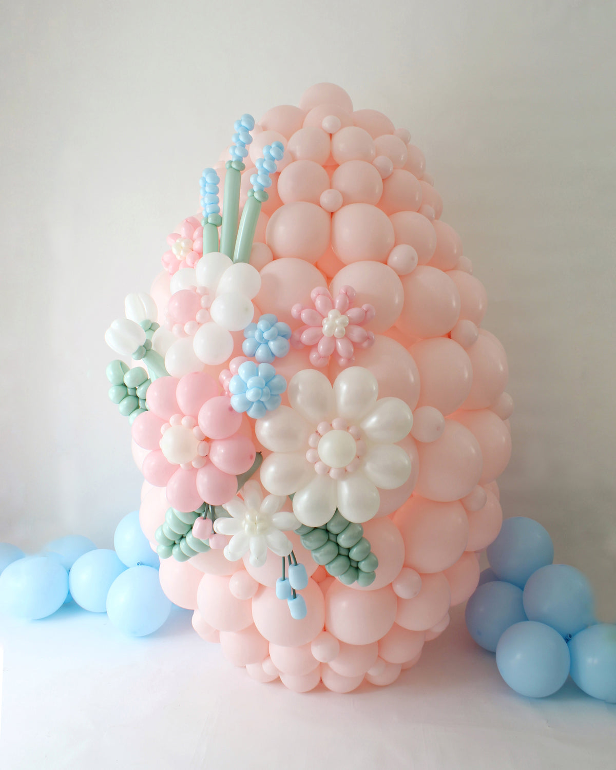 Large pastel pink balloon Easter egg decorated with 3D balloon flowers in soft shades of white, blue, and pink. Designed as part of an Easter balloon display, this eye-catching piece is surrounded by light blue balloon clusters, making it a perfect statement decor for spring celebrations or Easter-themed parties.