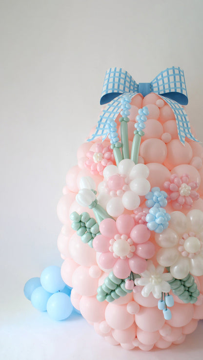 Large pastel pink balloon Easter egg decorated with 3D balloon flowers, greenery, and topped with an oversized blue gingham paper bow. Designed as a spring centerpiece or party backdrop, this balloon sculpture features soft Sempertex balloon tones in pink, white, mint, and blue—perfect for Easter events, photo ops, or Creative Heart Club members recreating the class tutorial.