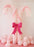 Two large candy cane columns made from pastel pink and white balloons stand side by side, each with a curved top and textured balloon pattern. A bold hot pink paper bow is placed at the center, creating a festive focal point. Soft pink balloons are scattered on the floor around the base, making the setup perfect for Christmas parties, kids’ events, and holiday photo backdrops.