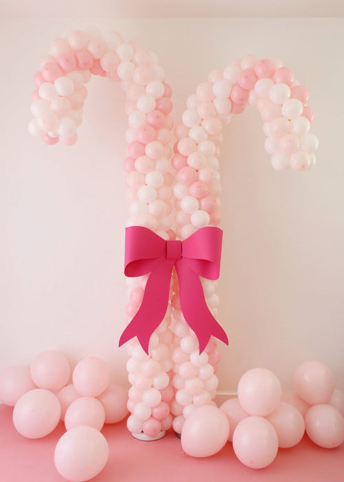 Two large candy cane columns made from pastel pink and white balloons stand side by side, each with a curved top and textured balloon pattern. A bold hot pink paper bow is placed at the center, creating a festive focal point. Soft pink balloons are scattered on the floor around the base, making the setup perfect for Christmas parties, kids’ events, and holiday photo backdrops.