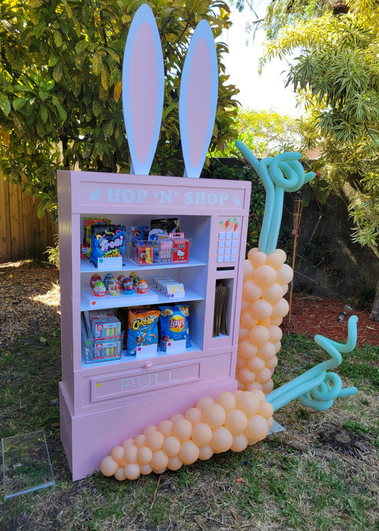 Pastel pink DIY foamboard vending machine prop decorated with large bunny ears and balloon carrots, labeled "Hop 'N' Shop." Filled with colorful candy and Easter-themed treats, this party prop is perfect for spring events, kids' parties, or Easter photo booths. Built using templates from the Foamboard Vending Machine Class, it showcases a creative use of foam board and balloon decor.