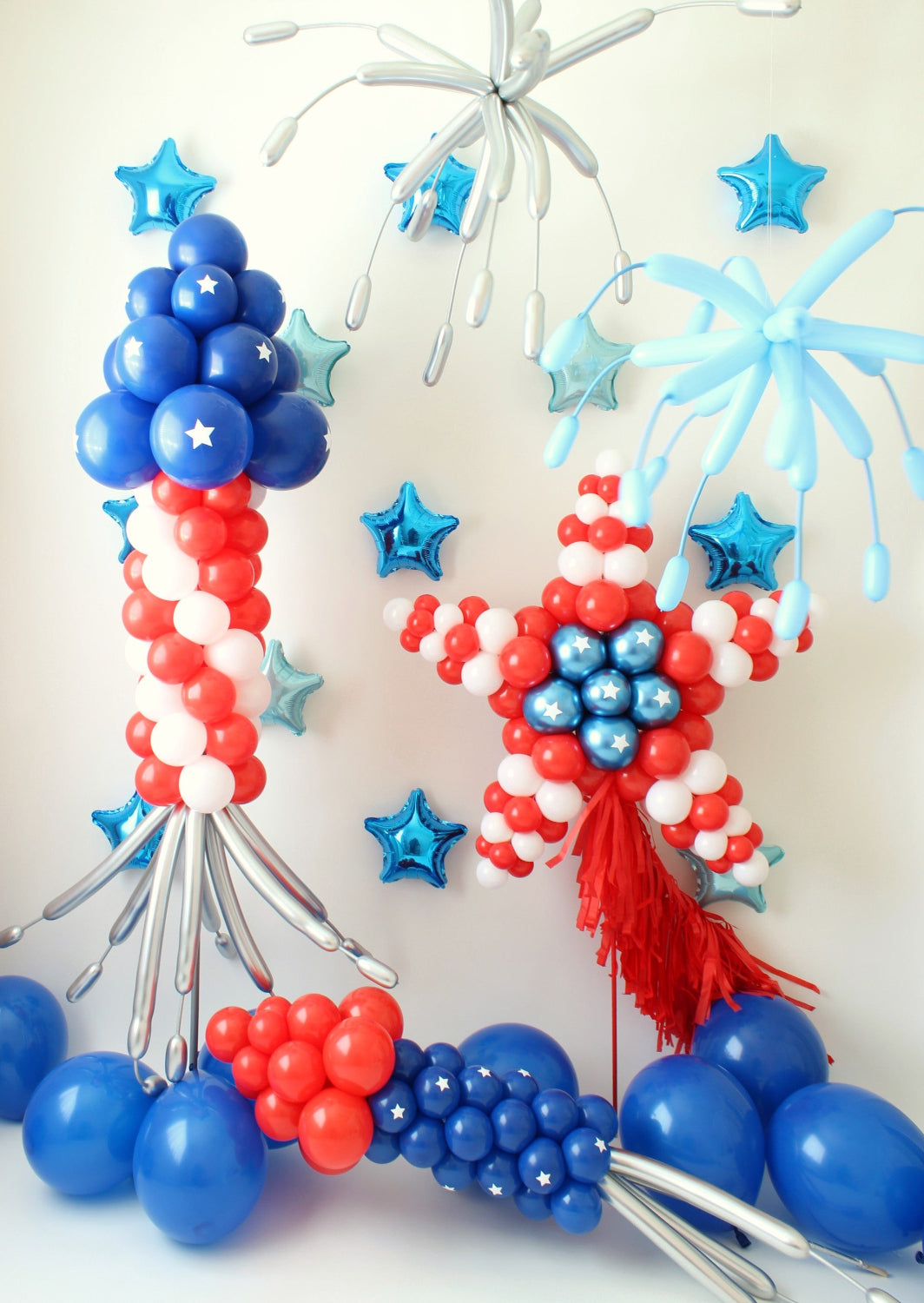 A vibrant patriotic balloon display featuring a striped bottle rocket column, a star-shaped balloon firework with fringe, and a small bottle rocket, surrounded by metallic blue star balloons and balloon fireworks bursting overhead. Designed by Lilly Jimenez of The Creative Heart Studio, this eye-catching setup is perfect for Fourth of July celebrations, parades, and patriotic-themed events. 🎆🎈