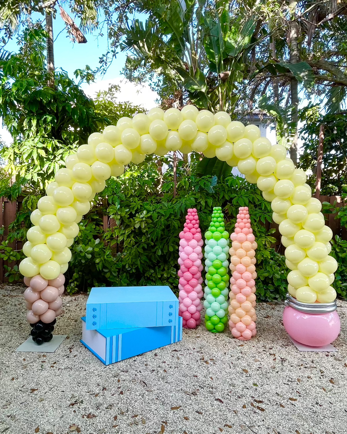 Outdoor setup featuring a pencil-shaped balloon arch paired with four crayon-style balloon columns in pink, white, green, and coral, plus a giant open book prop in blue. This colorful back-to-school balloon display is perfect for school events, teacher appreciation, and classroom photo booths. Designed for the Pencil Balloon Arch Tutorial.