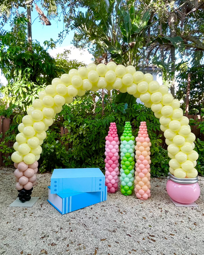 Outdoor setup featuring a pencil-shaped balloon arch paired with four crayon-style balloon columns in pink, white, green, and coral, plus a giant open book prop in blue. This colorful back-to-school balloon display is perfect for school events, teacher appreciation, and classroom photo booths. Designed for the Pencil Balloon Arch Tutorial.