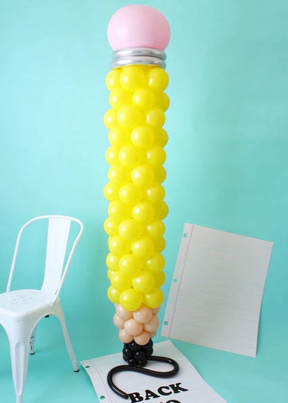 A tall pencil-shaped balloon column made with yellow balloons for the body, a pink balloon for the eraser, a silver band, and a brown and black base mimicking the sharpened tip. The column stands against a teal background, accompanied by a white chair and a large notebook paper prop, creating a fun and festive back-to-school display.