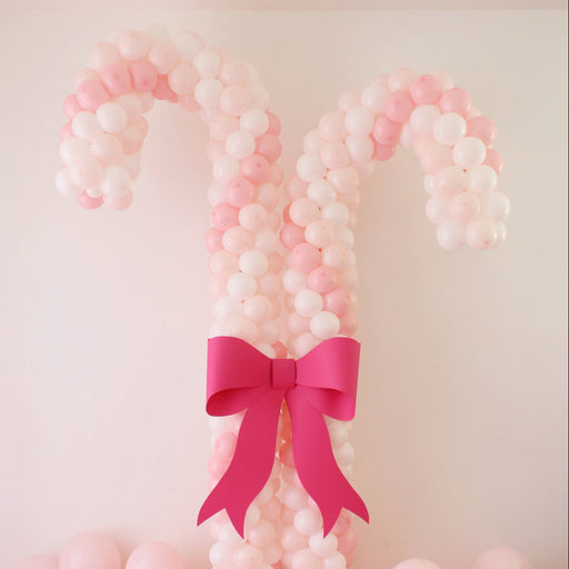Two oversized candy cane columns made entirely of pastel pink and white balloons stand side by side, styled with a smooth gradient effect from light to darker pink. A large hot pink paper bow is placed at the center where the two balloon candy canes cross, creating a cute holiday focal point perfect for Christmas parties, photo ops, and festive backdrops.