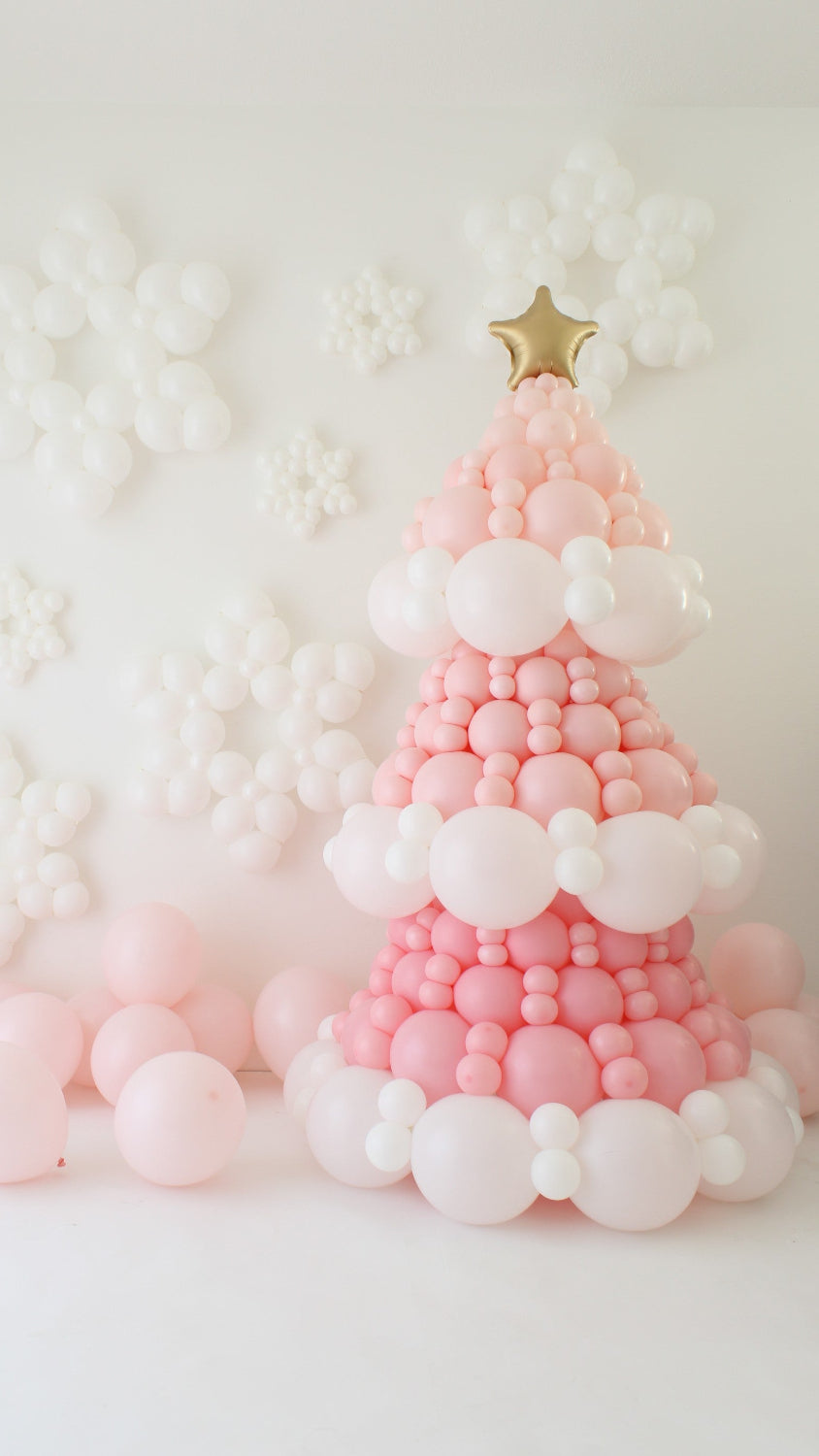 Pink balloon Christmas tree made with layered pink and white balloons, topped with a gold star balloon. White balloon snowflakes cover the wall behind it, with soft pastel pink balloons at the base creating a cute winter backdrop for holiday decor and Christmas party photo setups.