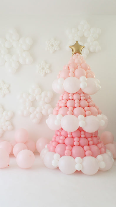 Pink balloon Christmas tree made with layered pink and white balloons, topped with a gold star balloon. White balloon snowflakes cover the wall behind it, with soft pastel pink balloons at the base creating a cute winter backdrop for holiday decor and Christmas party photo setups.