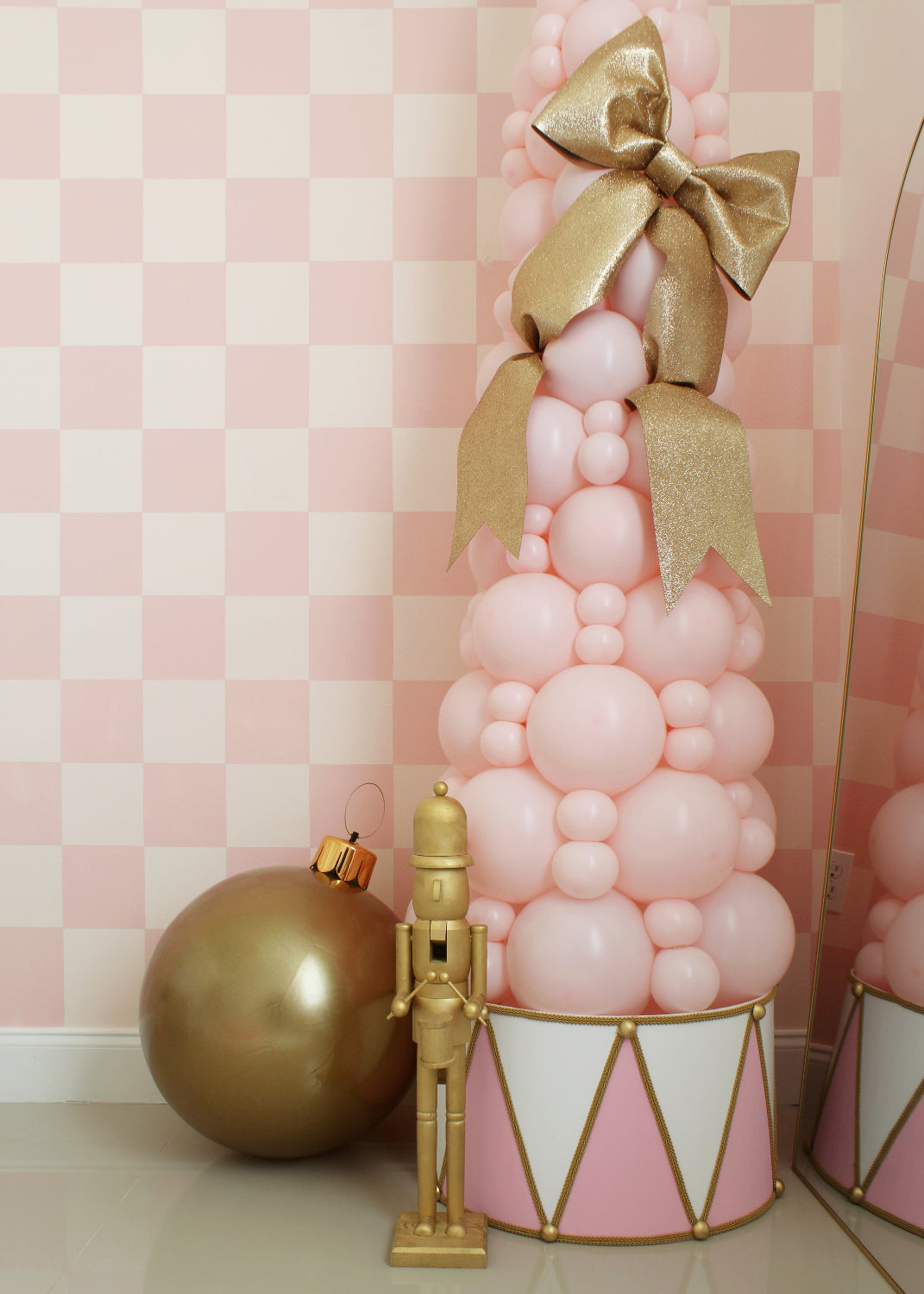 Whimsical Christmas display featuring a pastel pink balloon tree topped with a large sparkling gold bow. The tree is accented by a pink and white foam board drum base with gold trim, creating a festive and elegant foundation. Beside the tree, a gold nutcracker and oversized gold ornament add extra holiday charm. A magical and photo-worthy design ideal for Christmas parties, kids' events, or holiday window decor. Created by Lilly Jimenez of Creative Heart Studio.