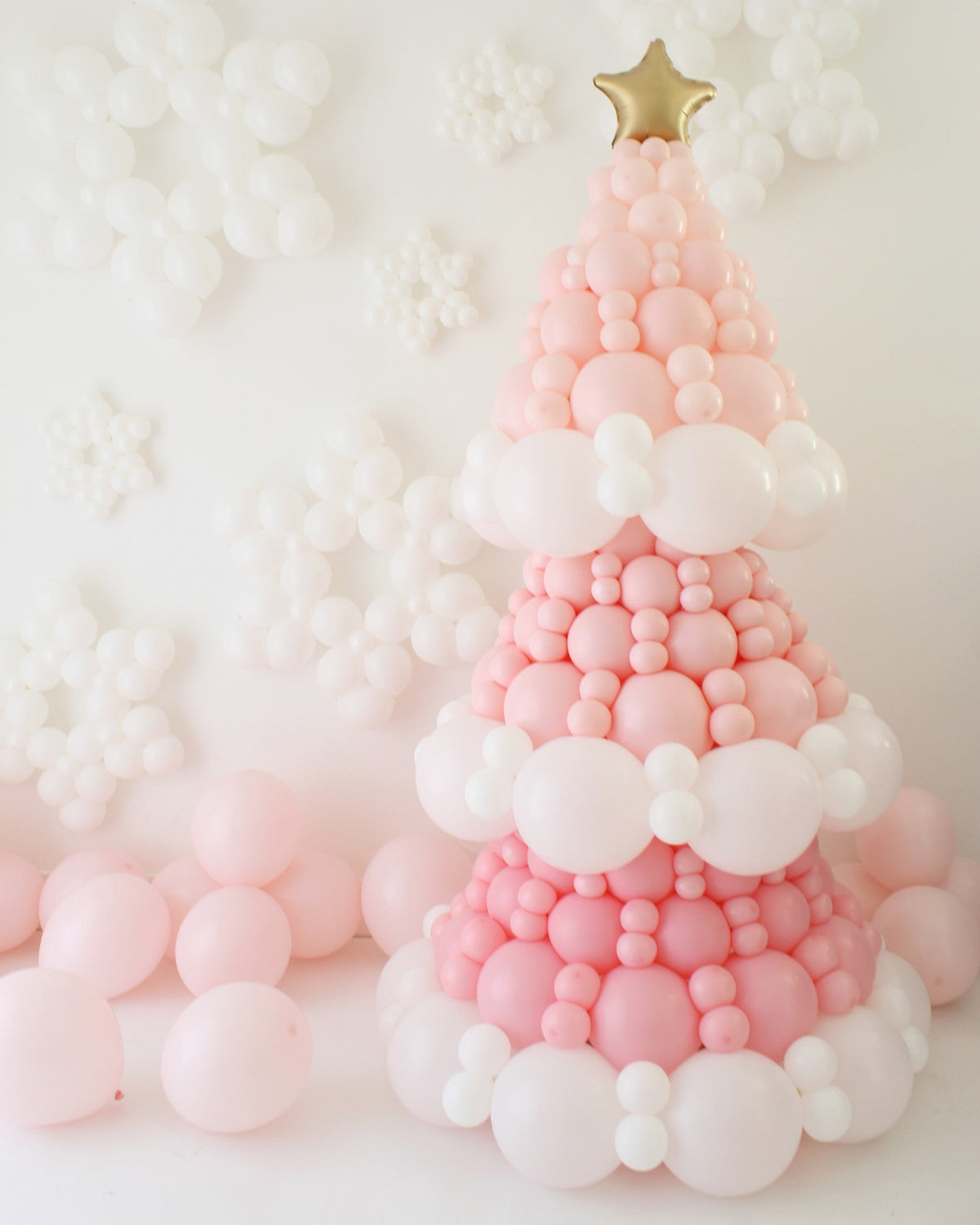 Pink balloon Christmas tree with a slightly pointed top made from layered pink and white balloons and finished with a gold star. White balloon snowflakes cover the wall behind it, and soft pastel pink balloons sit at the base. This whimsical holiday backdrop is the same design you can learn step by step inside the Whimsical Balloon Christmas Tree Class, ideal for Christmas installs and photo setups.