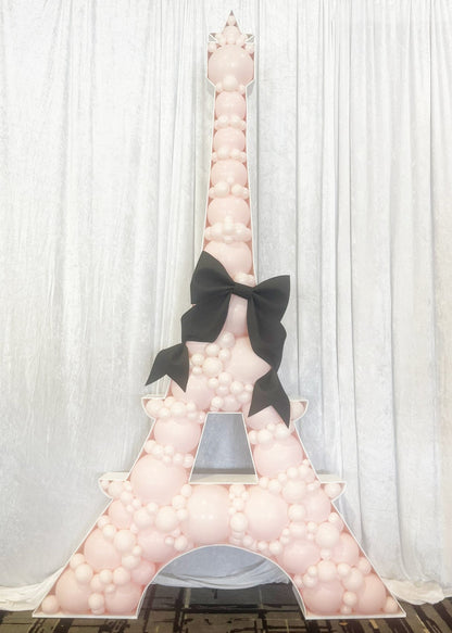 Eiffel Tower-shaped balloon mosaic filled with blush pink balloons in various sizes, topped with a large black bow at the center. The design stands against a white fabric curtain backdrop, creating an elegant and whimsical Paris-themed decoration. Ideal for bridal showers, birthday parties, or girly events, this setup showcases the finished look achievable using the Eiffel Tower Balloon Mosaic Template by Creative Heart Studio.