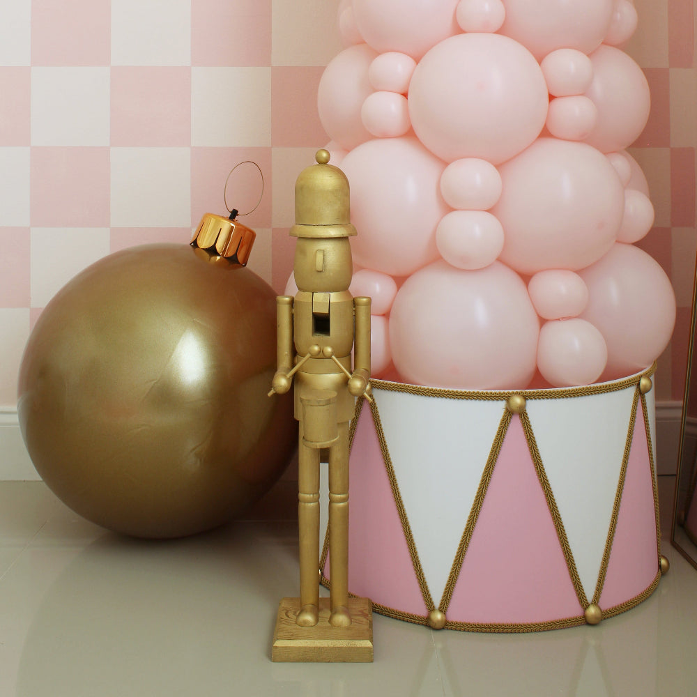 Elegant pink and white foam board Christmas drum with gold rope and trim detail sits around the base of a pastel pink balloon tree. A gold nutcracker and oversized gold ornament complete the festive scene. Designed by Lilly Jimenez, this charming DIY drum adds a whimsical and polished look to Christmas balloon decor, perfect for holiday events, window displays, or party backdrops. A standout piece that transforms your seasonal setup into a magical moment.