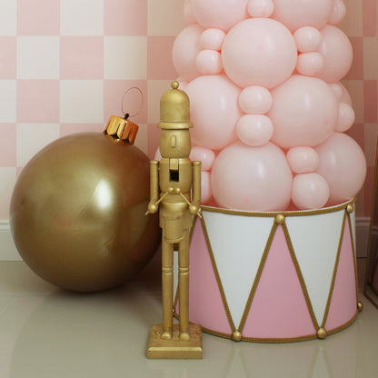 Elegant pink and white foam board Christmas drum with gold rope and trim detail sits around the base of a pastel pink balloon tree. A gold nutcracker and oversized gold ornament complete the festive scene. Designed by Lilly Jimenez, this charming DIY drum adds a whimsical and polished look to Christmas balloon decor, perfect for holiday events, window displays, or party backdrops. A standout piece that transforms your seasonal setup into a magical moment.