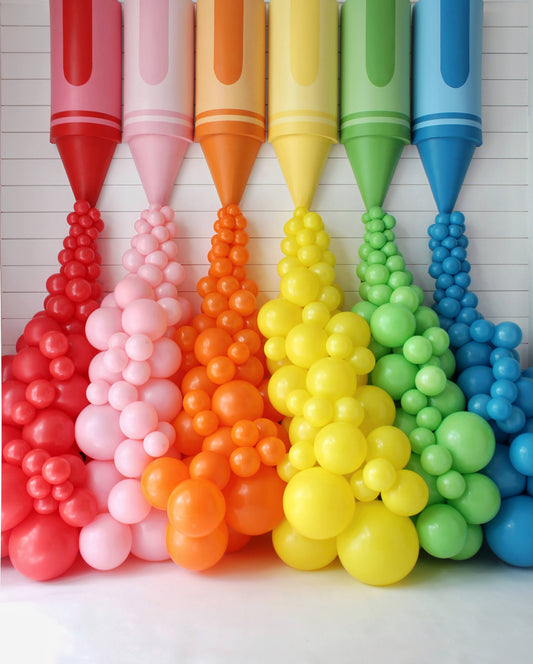 Oversized rainbow crayon props with matching balloon garlands spilling from each crayon tip, creating a vibrant, colorful classroom or party backdrop. Designed by Lilly Jimenez for her Crayon Prop Tutorial at Creative Heart Studio, this display is perfect for back-to-school events, photo ops, teacher appreciation setups, or any celebration needing a pop of playful creativity.