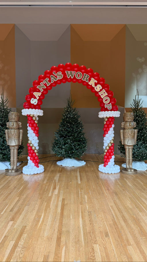 Santa’s Workshop red and white balloon arch with holiday lettering, candy cane style columns, Christmas trees, and nutcracker accents, ideal for festive photo backdrops, Santa events, and winter balloon decor displays.
