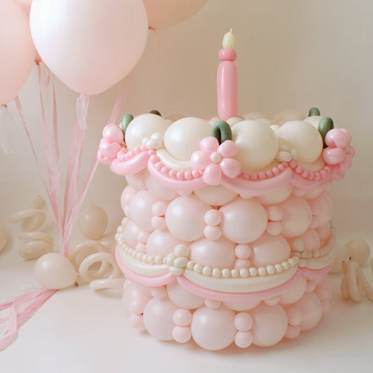 Pink and cream single-tier balloon cake with a pink candle, pastel balloon details, and soft pink helium balloons in the background.