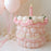 Pink and cream single-tier balloon cake with a pink candle, pastel balloon details, and soft pink helium balloons in the background.
