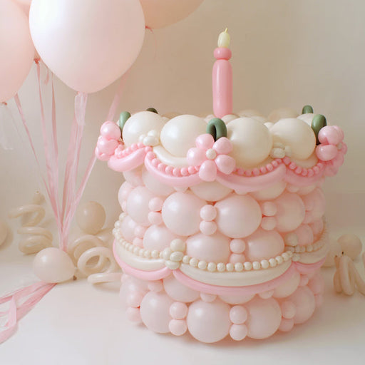 Pink and cream single-tier balloon cake with a pink candle, pastel balloon details, and soft pink helium balloons in the background.