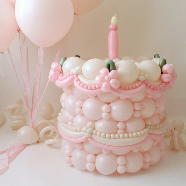 Pink and cream single-tier balloon cake with a pink candle, pastel balloon details, and soft pink helium balloons in the background.
