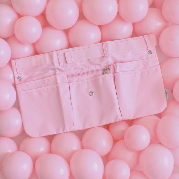 Solid light pink maker apron with six pockets, including a center zip pouch, belt loops, and grommets for tool attachments. Displayed on a background of pink balloons, this stylish and functional workbelt is designed for balloon artists, event decorators, and creative professionals. Its durable construction and adjustable fit make it perfect for holding pumps, scissors, and tools during events and installations.