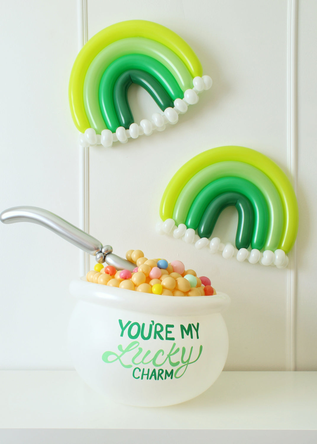 Festive balloon decor featuring a white cereal bowl balloon labeled “You’re My Lucky Charm,” filled with colorful balloon cereal and a silver spoon. Two green monochrome rainbow balloons with white balloon clouds are displayed above, creating a playful St. Patrick’s Day setup. Perfect for themed parties, festive photo ops, and part of Lilly Jimenez’s You’re My Lucky Charm Balloon Class.
