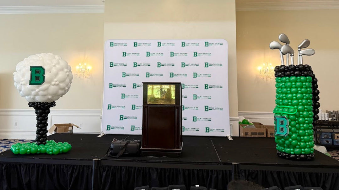 Stage balloon decor for a golf tournament featuring a balloon golf bag structure with silver golf clubs and a balloon golf ball on a tee. Displayed on an indoor event stage with a branded backdrop, this professional golf themed balloon decor is ideal for tournaments, corporate events, and award ceremonies by The Creative Heart Studio.
