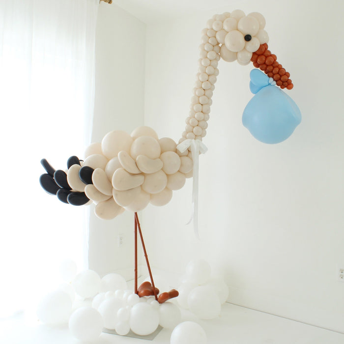 Neutral stork balloon sculpture holding a blue balloon bundle, designed as a welcome baby display for newborn arrivals, baby showers, nursery decor, and professional balloon installations.