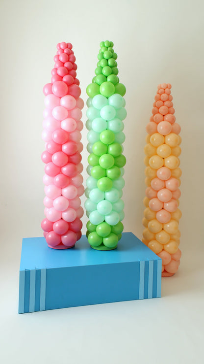 Three Balloon Crayon Columns in pink, green, and orange displayed on a large blue book prop. These colorful balloon sculptures are part of the Back to School Column Class, a tutorial series for creating school-themed balloon decor. Includes video lessons and printable balloon recipes, perfect for classroom events, teacher appreciation, and back to school photo setups.