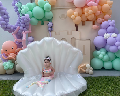 A toddler celebrates her 1st birthday dressed as a mermaid, sitting on an inflatable seashell chair in front of a DIY foam board Sand Castle Backdrop. The setup features pastel balloon garlands, coral, and sea creatures, all part of the Sand Castle Backdrop Class from The Creative Heart Studio—perfect for under-the-sea or beach-themed first birthday celebrations.
