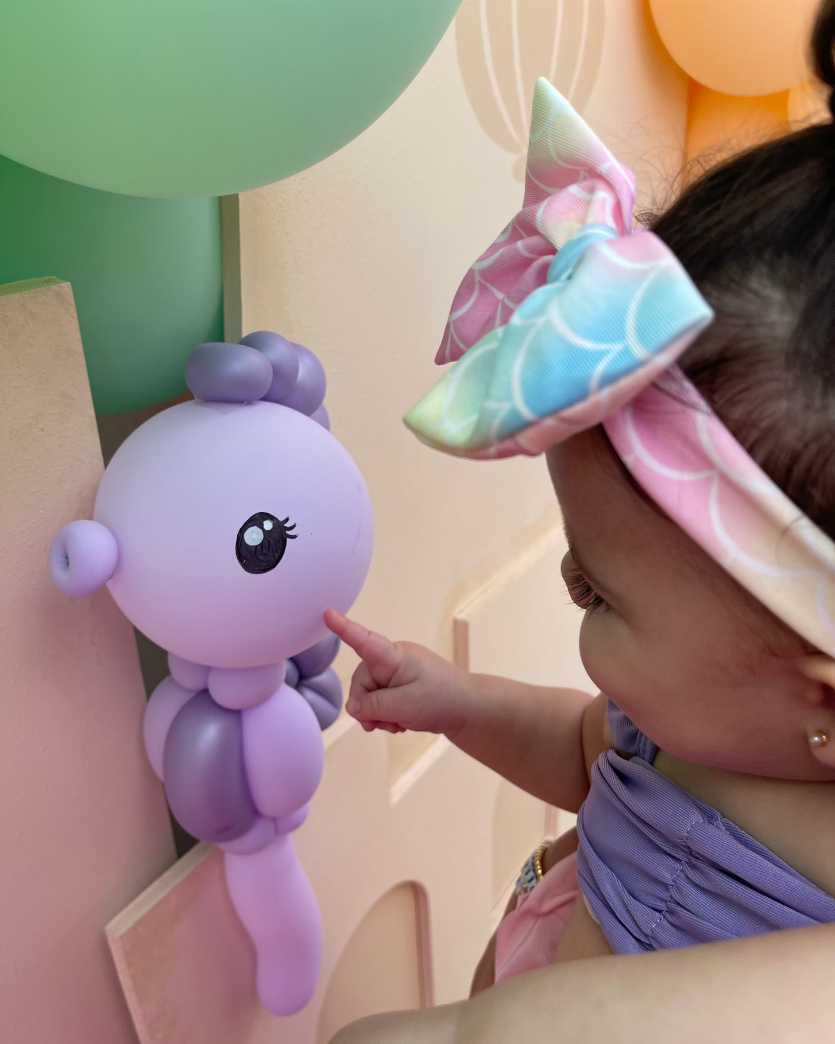 Close-up of a toddler gently touching a purple balloon seahorse attached to a foam board Sand Castle Backdrop. She wears a pastel mermaid bow and outfit while celebrating her first birthday at an under-the-sea themed party. The balloon sculpture and backdrop are part of the Sand Castle Backdrop Class by The Creative Heart Studio, designed to bring magical detail to birthday and ocean-themed events.

