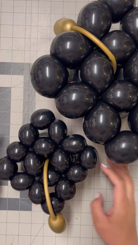 Two mini graduation caps made from black balloons are displayed side-by-side, each with a gold balloon tassel. One is smaller and one larger, perfect for candy cups, tabletop displays, or balloon arrangements for graduation celebrations. Ideal for balloon artists, party decorators, or DIYers creating fun graduation-themed decor.