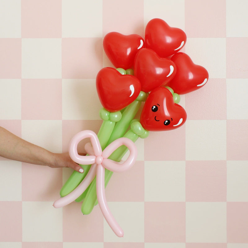 Balloon bouquet of red heart-shaped flowers with a smiling face, tied with a pink balloon bow, against a pink checkered background.