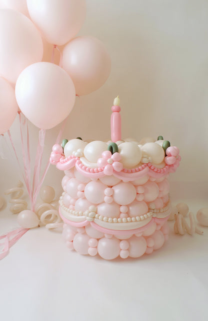 A vintage-inspired balloon cake featuring intricate pearl-like balloon details, delicate pink swirls, and floral accents, topped with a single balloon candle. Soft pink balloons in the background enhance the elegant aesthetic. Designed by Lilly Jimenez of The Creative Heart Studio, this charming balloon cake is perfect for weddings, birthdays, and sophisticated celebrations. Learn how to create this stunning piece in the Balloon Cake Class!