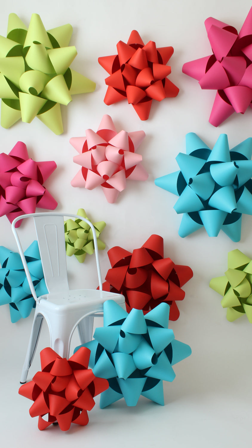 A vibrant display of oversized gift bows in red, pink, blue, and green arranged on a white wall. Some bows are mounted on the wall, while others rest on the floor beside a white metal chair, creating a playful and festive atmosphere perfect for event decor or holiday celebrations.