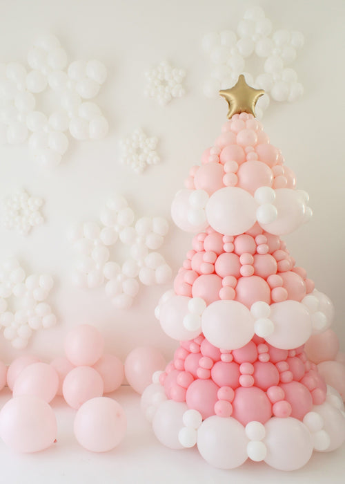 Pink balloon Christmas tree built with layered pink and white balloons and topped with a gold star. White balloon snowflakes decorate the wall behind it with pastel balloons gathered at the base. This holiday backdrop is the same design you can learn step by step inside the Whimsical Balloon Christmas Tree Class, perfect for balloon artists and Christmas installs.